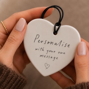 May include: A white heart-shaped ceramic ornament featuring the black printed text "Personalise with your own message" and a small hand-drawn heart icon. A black cord is looped through a hole at the top for hanging this decorative piece.