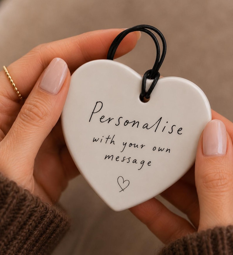 May include: A white heart-shaped ceramic ornament featuring the black printed text "Personalise with your own message" and a small hand-drawn heart icon. A black cord is looped through a hole at the top for hanging this decorative piece.