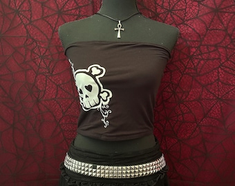 Puede incluir: Top de tubo negro con un gráfico de calavera blanca y un cinturón con tachuelas. La calavera tiene un ojo en forma de corazón. Un colgante de ankh plateado cuelga de una gargantilla negra. El atuendo se muestra en un maniquí negro.