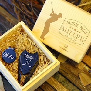 Könnte beinhalten: Eine Holzgeschenkbox mit Golf-Motiv. Sie enthält einen Golfballmarker und ein Pitchgabel, beide marineblau und mit "Miller" graviert. Der Deckel ist mit einer Golfersilhouette und dem Text "Groomsman Miller 09/28/22" graviert.