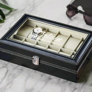 May include: Black leather watch box with a glass top and white stitching. The box has 10 compartments for watches and the text "Calvin Best Man" is printed on the inside of the lid.