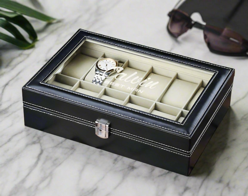 May include: Black leather watch box with a glass top and white stitching. The box has 10 compartments for watches and the text "Calvin Best Man" is printed on the inside of the lid.