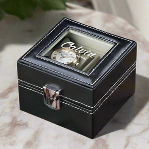 May include: Black leather watch box with a clear lid and white stitching. The box has a silver latch and the name "Calvin" is engraved on the lid.