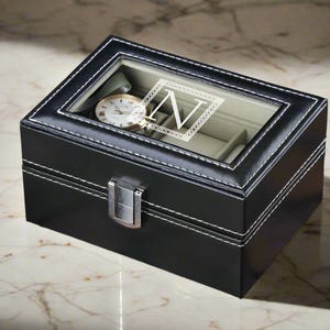 May include: Black leather watch box with a clear lid and a single watch inside. The box has a white stitched border and a silver metal latch. The lid has a white monogrammed "N" in a decorative frame.