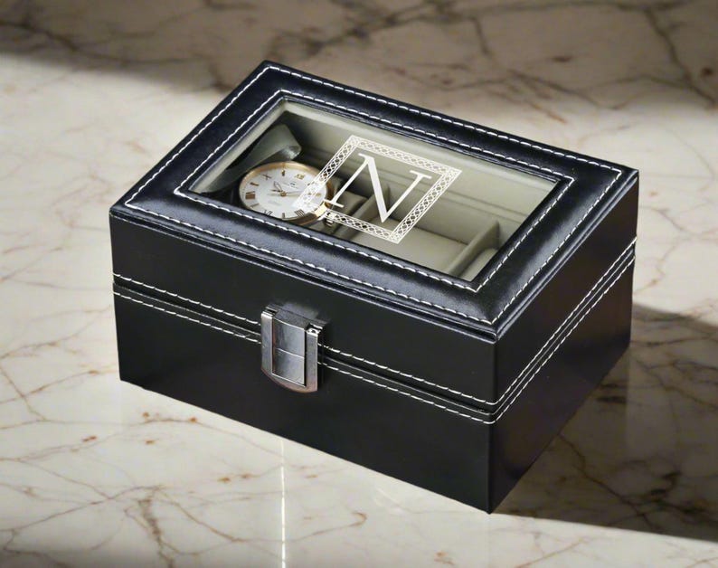 May include: Black leather watch box with a clear lid and a single watch inside. The box has a white stitched border and a silver metal latch. The lid has a white monogrammed "N" in a decorative frame.