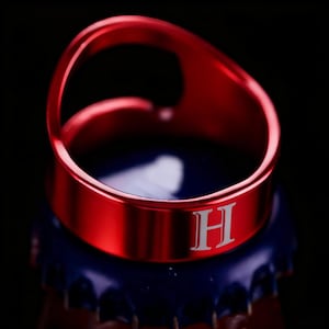 Peut inclure: Une bague ouvre-bouteille rouge avec la lettre "H" gravée sur le bandeau. La bague en métal est posée sur un bouchon de bouteille bleu, sur fond noir.