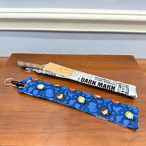 May include: Two wand holders, one with a blue fabric with a pattern of Harry Potter characters and the other with a white fabric with a newspaper print and the text "Dark Mark".