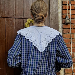 May include: A blue, green, and white plaid blouse with a white, eyelet lace collar. The blouse has long sleeves and a ruffled hem. The person is facing away from the camera, with their hair in a bun. The background includes a wooden door and a brick wall.