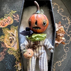 May include: A small pumpkin-headed doll wearing a striped outfit and a green collar, holding a smaller pumpkin in its hand. The doll is displayed in a black coffin-shaped box.