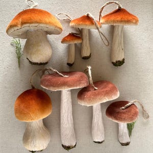 May include: A collection of decorative mushroom ornaments with orange and brown velvet caps and white stems. Each mushroom has a twine loop for hanging. The ornaments vary in size and shape, perfect for seasonal decor.