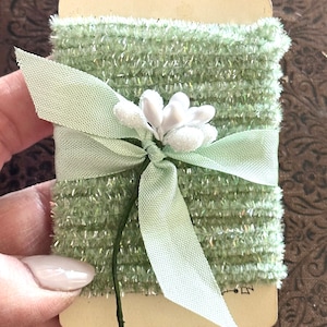 May include: A roll of green sparkly ribbon with a white flower tied in a bow. The ribbon is on a card with the word "Z" written on it.