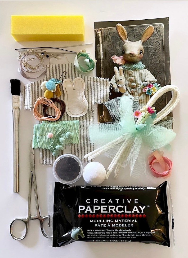 May include: A craft kit for making a bunny doll. The kit includes white paper clay, a small container of black paint, a white pom-pom, a pair of scissors, a paintbrush, a piece of white fabric with bunny ears, a piece of striped fabric, a piece of green fabric, a piece of white tulle, a piece of pink ribbon, a piece of white pipe cleaner, a piece of yellow sponge, and a piece of white floral wire with a green bow.