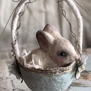 May include: A small, white bunny sits inside a blue and white egg-shaped basket with a white and silver trim. The basket is hanging from a white pipe cleaner with a silver wire wrapped around it.