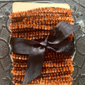 May include: A bundle of orange and black fuzzy trim, tied with a black satin ribbon. The trim is wrapped around a cream-colored card. The overall aesthetic is vintage and decorative.
