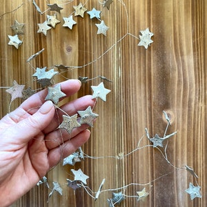 May include: A string of silver and gold star-shaped ornaments hanging on a twine string. The stars are made of a textured material and have a rustic look.