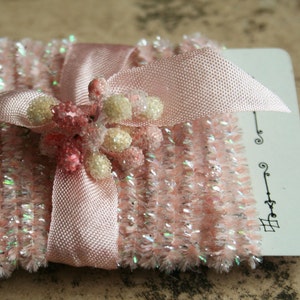 May include: A pink, textured, decorative item with a satin ribbon and floral accents. The item is embellished with iridescent glitter and small, white, and pink floral clusters. A white card with black decorative lines is partially visible.