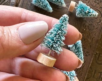 May include: Miniature teal bottle brush Christmas trees with white frosted tips and wooden bases. Several trees are scattered on a vintage book. One tree is held in a hand, showcasing the size and detail of the holiday decor.