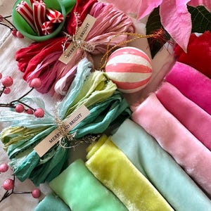 May include: A festive arrangement featuring a green cup filled with red and white striped candies, colorful fabric bundles, a striped ornament, and a pink poinsettia. The fabric bundles have tags that read "MERRY AND BRIGHT".