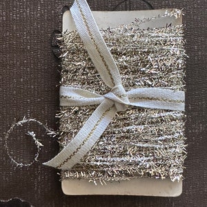 May include: A spool of silver tinsel ribbon with a white ribbon tied around it. The ribbon is on a white card.