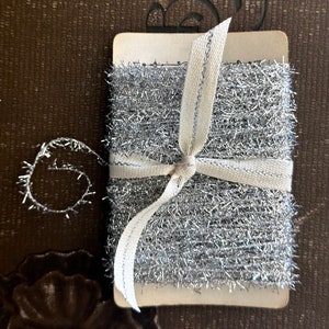 May include: A roll of silver tinsel ribbon tied with a white ribbon. The tinsel is a sparkly, metallic material.