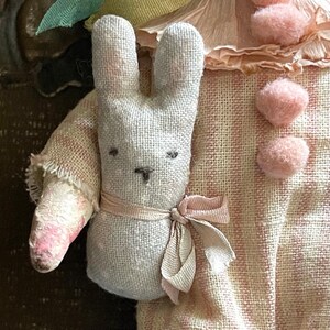 May include: A handmade doll wearing a pink and white striped dress with a white bunny attached to its arm. The doll has a pink flower on its head and pink pom-poms on its dress.
