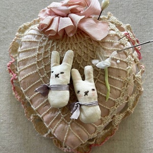 May include: A heart-shaped pincushion with crocheted lace trim, featuring a pink satin bow and two white fabric bunny figurines with gray ribbon accents.