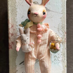 May include: A handmade pink and white bunny doll wearing a pink and white striped outfit, holding a small white bunny doll and a basket with a yellow chick inside. The doll is standing on a canvas background with a vintage design.