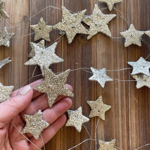May include: A silver glitter star garland with a string of silver stars connected by a thin wire. The garland is hanging on a wooden surface.