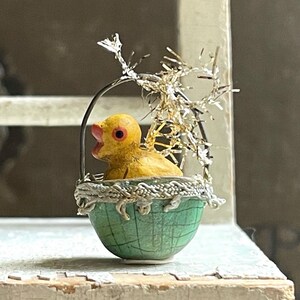 May include: A yellow chick figurine sits inside a green and white egg-shaped basket with a silver tinsel bow. The chick has a red beak and eyes.