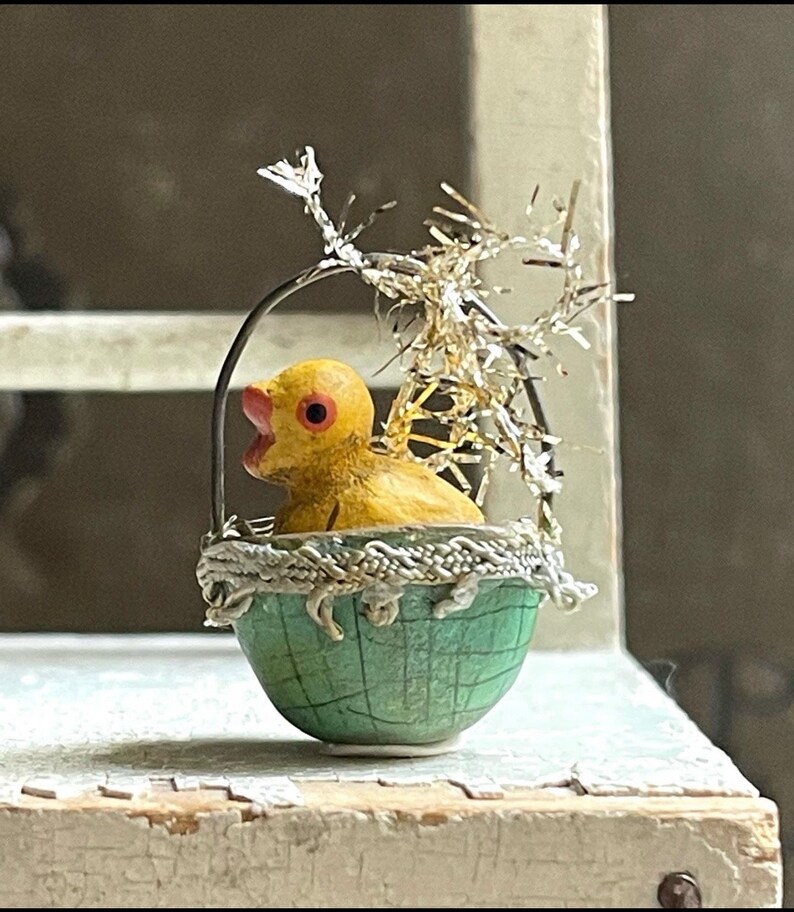 May include: A yellow chick figurine sits inside a green and white egg-shaped basket with a silver tinsel bow. The chick has a red beak and eyes.