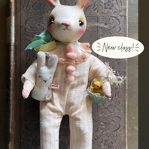 May include: A white bunny doll wearing a pink and white striped outfit, holding a small white bunny doll and a basket with a yellow chick inside. The doll has pink cheeks and black eyes. The background is a brown book with a floral design. The text "New class!" is written on a white speech bubble.