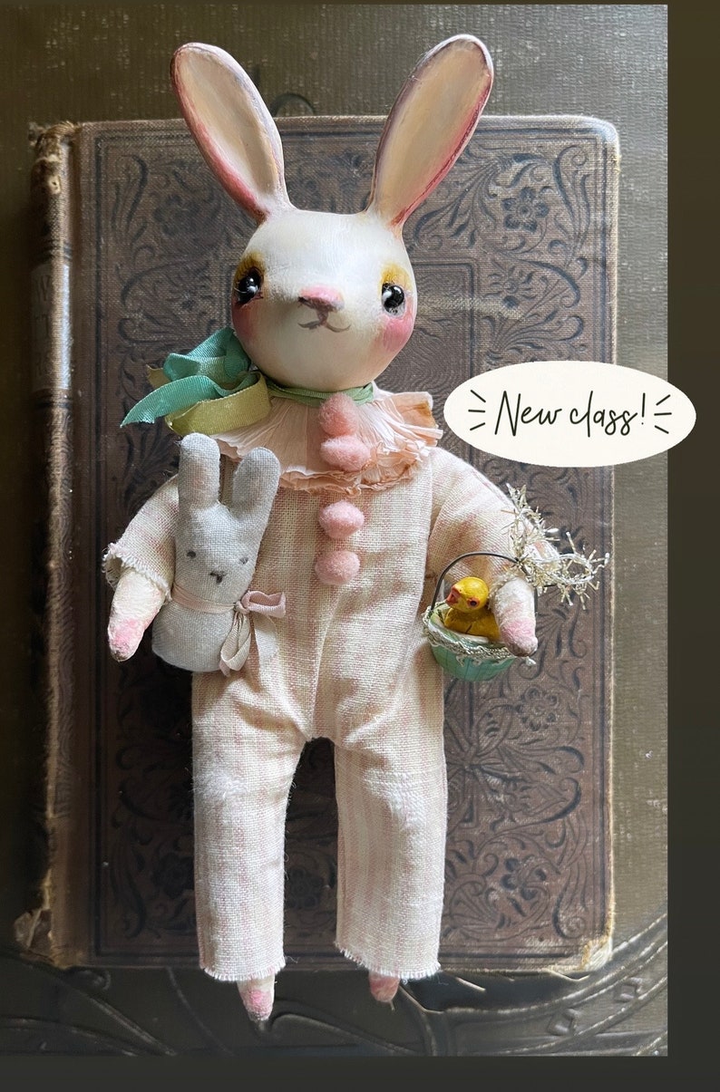 May include: A white bunny doll wearing a pink and white striped outfit, holding a small white bunny doll and a basket with a yellow chick inside. The doll has pink cheeks and black eyes. The background is a brown book with a floral design. The text "New class!" is written on a white speech bubble.