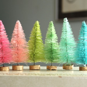 May include: Six miniature artificial Christmas trees in a gradient of pink, green and blue colours. The trees are on a wooden surface.