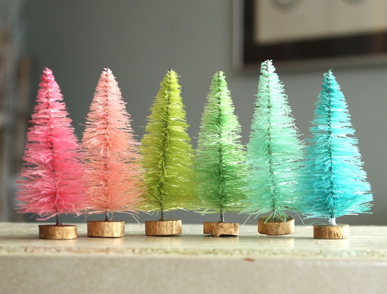 May include: Six miniature artificial Christmas trees in a gradient of pink, green and blue colours. The trees are on a wooden surface.