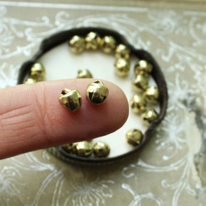 May include: A close-up of two small gold jingle bells. The bells are on a finger, and a container of more bells is in the background.
