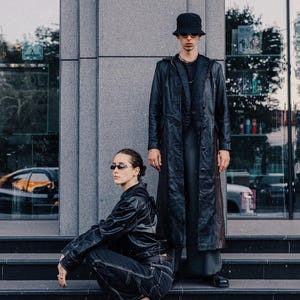 Puede incluir: Dos personas vestidas de negro posan en escalones frente a un edificio. Una persona está en cuclillas, con una chaqueta negra, vaqueros y tacones. La otra persona está de pie, con un abrigo largo negro, sombrero y gafas de sol.