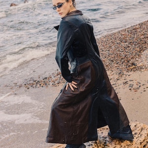 Puede incluir: Un abrigo largo de cuero negro con un panel marrón en la parte inferior. El abrigo se lleva con botas negras. La persona está de pie sobre una roca cerca del mar, usando gafas de sol.