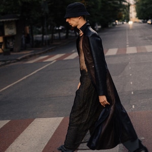 Puede incluir: Una persona con un sombrero de pescador negro, un abrigo largo negro y pantalones negros cruzando un paso de cebra. La persona también lleva zapatos negros. El fondo incluye árboles y una calle.