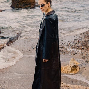 Puede incluir: Una persona con un abrigo largo de cuero marrón oscuro con un cuello marrón en contraste, de pie en una playa rocosa. El abrigo llega hasta los tobillos y tiene un diseño elegante. La persona también lleva gafas de sol.