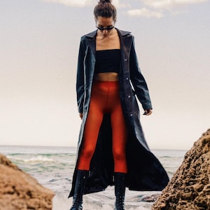 Puede incluir: Una persona con un abrigo largo de cuero negro, un top negro y leggings naranjas, de pie sobre una roca cerca del mar. La persona también lleva botas negras y gafas de sol. El cielo está nublado.
