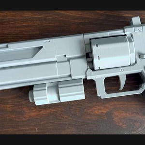 Peut inclure: Un jouet pistolet en plastique gris avec un canon, une poignée et un chargeur. Le pistolet a un design futuriste.