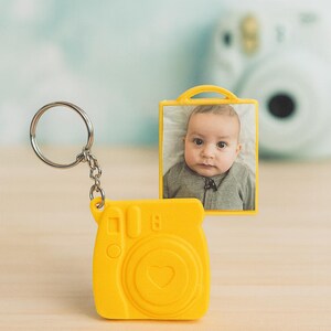 May include: A yellow camera-shaped keyring with a silver ring and chain. A small photo of a baby is in a yellow frame. A white camera is blurred in the background. The keyring has a heart shape.