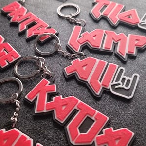 Könnte beinhalten: Eine Sammlung personalisierter Schlüsselanhänger mit Namen wie Britny, Tila, Ali und Katja in einer kräftigen, roten, 3D-gedruckten Schrift mit schwarzen Umrandungen. Jeder Schlüsselanhänger verfügt über einen silbernen Metallring und eine Kette.