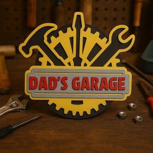 Puede incluir: Un letrero decorativo con el texto "DAD'S GARAGE" en letras rojas. El letrero presenta una forma de engranaje negro con herramientas amarillas, incluyendo un martillo, una llave y un destornillador. El letrero está sobre una superficie de madera.