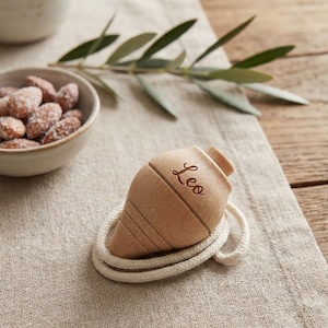 May include: A wooden spinning top with the name "Leo" engraved on its side, resting on a beige linen surface. A white string is wrapped around the top. A small bowl of sugared almonds and olive branches are in the background.