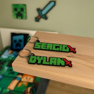Könnte beinhalten: Zwei Schlüsselanhänger mit den Namen "SERGIO" und "DYLAN" in grünen Blockbuchstaben mit rotem Schwertakzent. Die Schlüsselanhänger liegen auf einer Holzoberfläche, mit Minecraft-Wanddekoration im Hintergrund.