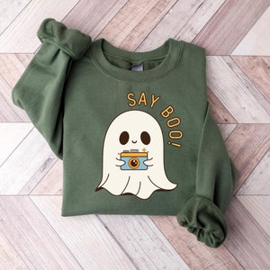Puede incluir: Sudadera verde con un fantasma blanco sosteniendo una cámara y el texto "Say Boo!" impreso en ella.