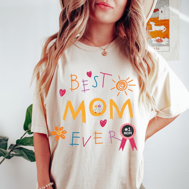 May include: A white t-shirt with the text "BEST MOM EVER" in colorful, childlike handwriting. The shirt also features a sun, flowers, and a pink ribbon with a badge that says "#1 MOM".