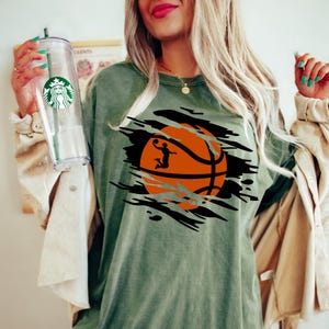 May include: Green t-shirt with a black and orange basketball graphic. The basketball is ripped open to reveal a silhouette of a basketball player.