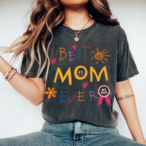 May include: A dark gray t-shirt with a colorful graphic that says "Best Mom Ever" with a sun, flowers, and hearts. The shirt also has a pink ribbon with a badge that says "#1 Mom".